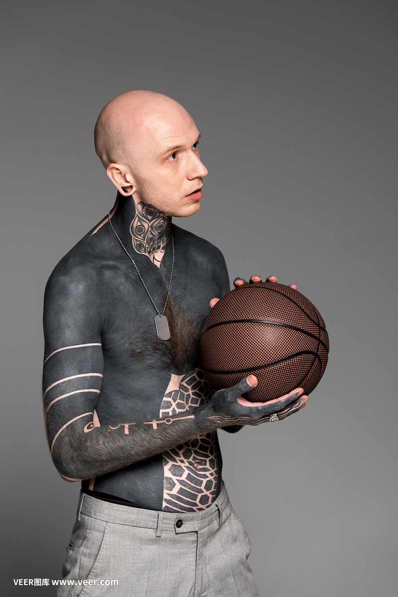 赤裸上身的光头纹身男子抱着篮球,孤立地看着灰色